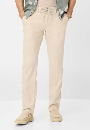 Pantalon en lin beige avec une coupe décontractée, taille élastique avec cordon de serrage et poches latérales, associé à des chaussures béiges à enfilage et une chemise à motifs.