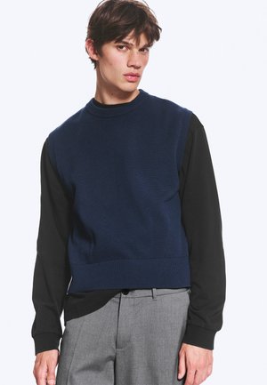 Jeune homme portant un pull sans manches bleu marine par-dessus une chemise noire à manches longues et un pantalon gris, posant devant un fond uni.