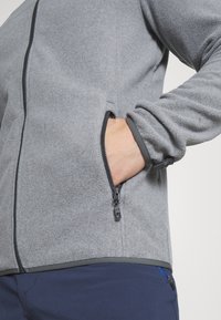 Hallhalli fleece-jakk, millel on tõmblukuga esiosa ja taskud. Omab elastseid mansette ja kontrastset äärt. Sile tekstuur, praktiline disain.