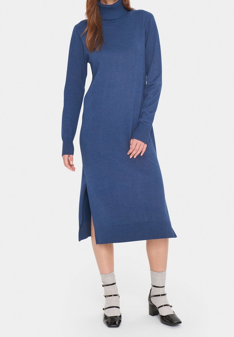 Robe à col roulé tricotée bleu foncé avec manches longues, fente latérale et poignets côtelés ; associée à des chaussettes grises et des chaussures à talons blocs noires.