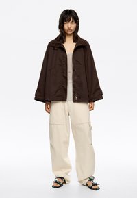 Mujer vestida con una chaqueta suelta de color marrón oscuro sobre una blusa transparente, pantalones anchos beige y sandalias de punta abierta con tiras decorativas sobre un fondo blanco.