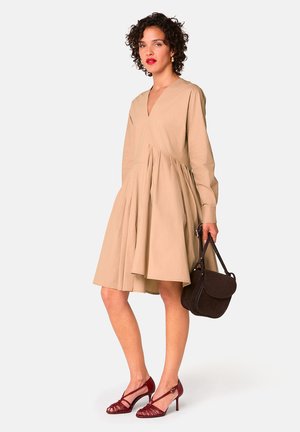 Beiges Langarmkleid mit V-Ausschnitt und plissiertem Rock. Das Model hält eine braune Tasche und trägt rote Riemchensandaletten. Glatte Textur.