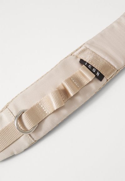 Beige fabric strap with a smooth texture, featuring a metal D-ring and layered fastening. Black tag with the word "ICON" visible.