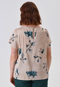 Frau mit schulterlangem, welligem braunem Haar, die eine beigefarbene Bluse mit blauen Blumenmustern trägt, von hinten vor einem schlichten Hintergrund betrachtet.