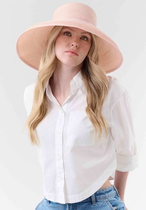 Femme aux longs cheveux blonds portant un chapeau rose à large bord, une chemise blanche à boutons et un jean bleu, face à l'avant avec une expression neutre.
