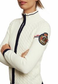 Vit stickad zip-up tröja med kabelmönster, marinblå detaljer och ett runt broderat emblem på vänster ärm.