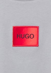 Un textil gris con una textura suave presenta un parche rectangular rojo que muestra "HUGO" en fuente negra, cosido en la esquina superior izquierda.