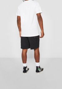 Valkoinen puuvillainen t-paita, rennolla istuvuudella, yhdistetty mustiin cargo-shortsseihin, joissa on taskuja. Mustat korkeavartiset kengät värikkäillä paloilla ja kuvioilla.