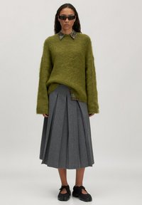 Olivengrüner, fusseliger Pullover mit lockerer Passform, kombiniert mit einem gefalteten grauen Rock und schwarzen Slippern. Enthält ein kariertes Kragen-Detail.