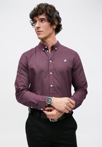 Bordó hosszú ujjú ing gombos gallérral, fehér logóval a mellkasán és gombos mandzsettával. Fekete nadrággal és karórával viselve.