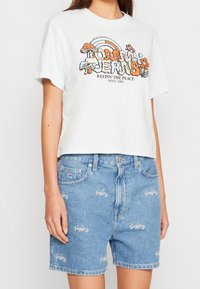 Žena v bílé grafické krátké tričko s houbami a duhou, spárované s modrými denimovými šortkami s vysokým pasem vyšívanými logy.