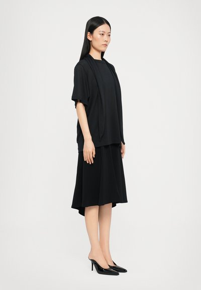 Outfit nero a strati composto da una blusa corta e morbida a maniche corte e una gonna che arriva al ginocchio. Tessuto liscio, design minimalista e décolleté a punta.
