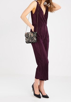 Femme portant une combinaison sans manches plissée de couleur pourpre foncé, des talons hauts noirs et un sac à bandoulière noir brodé de motifs floraux.