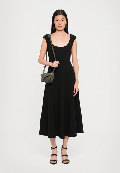 Robe noire sans manches avec encolure dégagée et jupe évasée, associée à des sandales noires à lanières et à un sac bandoulière vert olive.