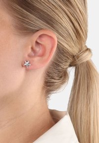 Primer plano de la oreja de una persona con un arete de estrella plateada, cabello rubio recogido en una coleta y un cuello de camisa blanca visible.