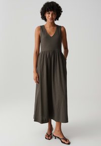 Woman with curly hair wearing a sleeveless dark olive maxi dress and black heeled sandals, standing with one hand in pocket.
