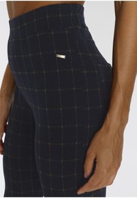Marineblaue Leggings mit einem Gittermuster aus goldenen Fäden, featuring einem hohen Taillenbund und einem kleinen metallischen Logo-Akzent an der Seite.