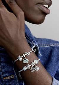Dvoje strieborných náramkov s príveskami, ktoré obsahujú rôzne charms: sovičku, srdce a ruky, s tyrkysovými akcentmi na zápästí proti džínsovine.