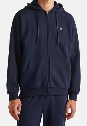 Mann trägt einen marineblauen Zip-Hoodie mit Fronttaschen und kleinem weißem Logo auf der linken Brust, kombiniert mit passenden marineblauen Hosen.
