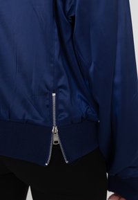 Marineblaue Bomberjacke aus glänzendem Material, mit einem silbernen Reißverschluss auf der Rückseite und gerippten Bündchen, mit einer glatten Textur und tailliertem Design.