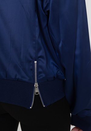 Marineblauwe satijnen bomberjack met geribbelde manchetten en een zilveren ritssluiting aan de achterkant, met een gladde textuur en reflecterend oppervlak.