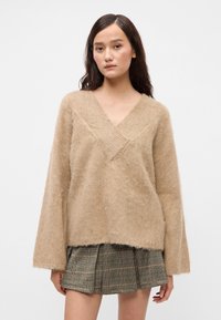 Jovem mulher com cabelo longo e escuro, vestindo um suéter bege de gola em V felpudo e uma saia plissada em padrão xadrez marrom, de pé em frente a um fundo branco liso.