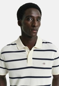 Crèmekleurige polo met marineblauwe strepen, korte mouwen en een kraag met drie knopen. Bevat een klein geborduurd logo op de borst.