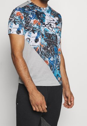 Mand iført kortærmet t-shirt med abstrakt mønster af blå, sort, hvid og orange malingstænk samt gråt diagonalt panel, kombineret med sorte bukser.