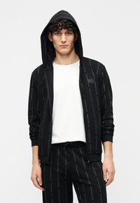 Sudadera con capucha negra con cierre, con un patrón repetitivo de logotipos. Está hecha de tela suave, con mangas largas y puños acanalados.