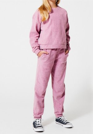 Personne debout portant un sweat-shirt en polaire rose et un pantalon assorti, les mains dans les poches, avec des baskets basses noires à lacets blancs.