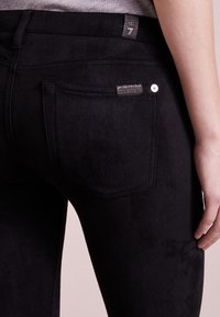Jeans noirs en tissu doux avec une texture semblable à du daim. Les caractéristiques comprennent des poches arrière, un bouton métallique et une étiquette en tissu avec la marque.