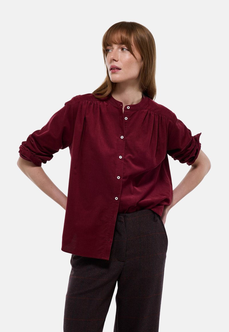 Chemise bordeaux à manches longues avec un col montant, fermeture par boutons à l'avant et détails froncés aux épaules. Tissu doux avec une texture lisse.