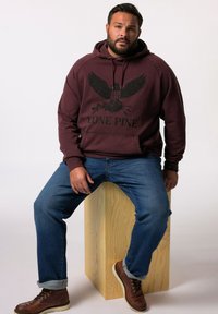 Bordeaux hoodie met een arendontwerp en de tekst "LONE PINE", gecombineerd met blauwe jeans en bruine laarzen, zittend op een houten blok.