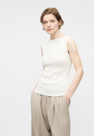 Νεαρή γυναίκα με κοντά μαλλιά, που φοράει αμάνικο λευκό τοπ με υφή και φαρδιά μπεζ παντελόνια, στέκεται με τα χέρια στις τσέπες.