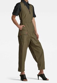 Kvinna iklädd en olivgrön ärmlös jumpsuit med frontdragkedja, sidofickor, svart kortärmad skjorta och svarta högklackade mule-skor.