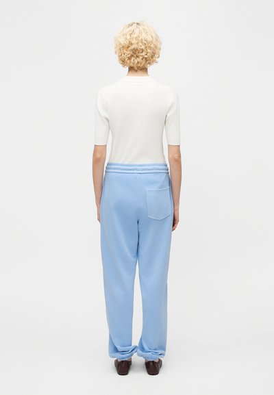 Άτομο με κοντά σγουρά ξανθά μαλλιά, που φορεί λευκό κοντομάνικο μπλουζάκι και φαρδιά ανοιχτό μπλε παντελόνια, από πίσω με φόντο ένα απλό φόντο.