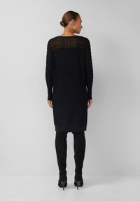 Femme avec les cheveux relevés en chignon, portant une robe noire à manches longues en côtelé et des bottes noires en daim à talons montant jusqu'au genou, se tenant devant un fond gris uni.