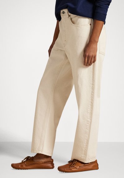 Μπεζ τζιν από denim με άνετη εφαρμογή, διαθέτοντας αντιθετικό ράψιμο και κλείσιμο με κουμπί, σε συνδυασμό με καφέ δερμάτινα παπούτσια τύπου slip-on.