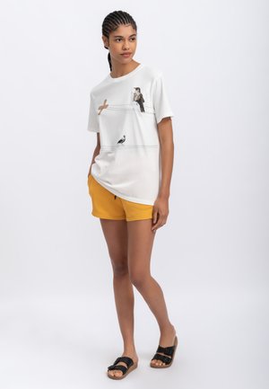Witte katoenen t-shirt met vogelafbeeldingen op een draad, gecombineerd met gele shorts. Het model draagt zwarte sliders.