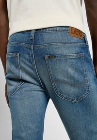 Niebieskie dżinsy mają lekko wyblakłe wykończenie, klasyczny pięciokomorowy design oraz brązową skórzaną naszywkę z logo Lee na tylnym pasku.