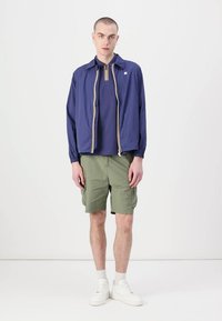 Marineblå lett zip-jakke med gullavskjæringer, brukt over en mørkeblå polo og grønne shorts. Modellen har på seg hvite sneakers og sokker.