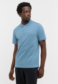 Jasnoniebieska koszulka polo o teksturowym wykończeniu, z trzema guzikami przy kołnierzy i małym logo na piersi. Połączona z ciemnymi spodniami.