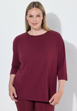 Vrouw met steil blond haar, gekleed in een donkerrood topje met driekwart mouwen en bijpassende broek, staand tegen een witte achtergrond.