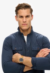 Camisa azul marino de botones con cuello alto y dos bolsillos en el pecho, combinada con un reloj de correa de cuero marrón con una carátula oscura.