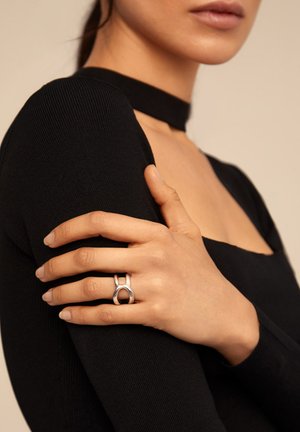 Bague en argent avec un design découpé portée sur une main contre un haut noir en côte. Le teint de la peau est visible, avec une finition naturelle des ongles.