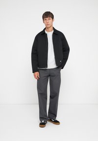Black, zip-up jacket with a fold-down collar, paired with gray trousers. The model wears black sneakers with brown accents. Smooth fabric texture.