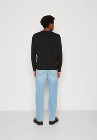 Camisa negra de manga larga con cuello redondo, combinada con jeans de pierna recta en azul claro y zapatos negros. Etiqueta amarilla en la parte inferior izquierda de la camisa.