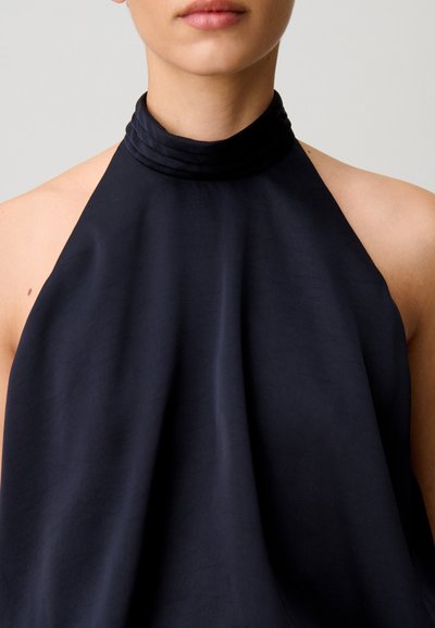 Donna che indossa un top nero smanicato a collo alto a coste, mostrata dal collo alla parte superiore del busto su uno sfondo semplice.