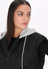 Gilet senza maniche nera trapuntata con cappuccio grigio, chiusura con zip e motivo testurizzato. Indossato sopra una maglia nera a maniche lunghe, evidenziando un design informale.