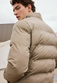 Jachetă puffer bej cu un design matlasat, având un guler înalt și o textură moale, netedă. Mânecele sunt umplute pentru căldură.